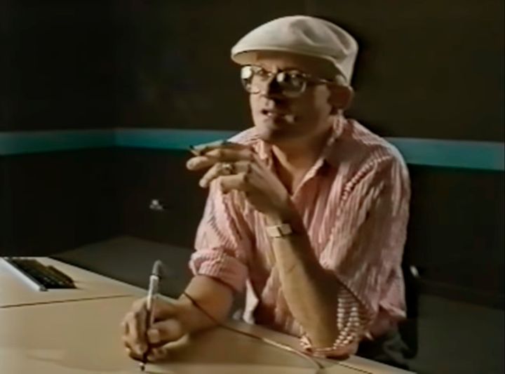 Screenshot from a video called "Painting with Light." David Hockney is around 49 years old and sitting at a large plastic desk with a metal stylus that is connected to the computer off-screen with a wire. He's wearing a white flat-cap and a light-weight pink and white striped shirt. Hockney is deep in focus, glossy-eyed with both elbows on the table, a cigarette casually in one hand.